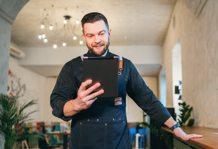 Smiling small business owner dressed in a black chef uniform with an apron using digital tablet in his cozy restaurant hall. Successful people, hard work, consumer cafes, restaurants industry concept.の写真素材