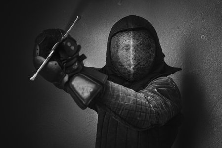 Portrait of a man dressed in black historical fencing armor and fencing protective helmet mask with a long medieval sharp sword prepared for aggressive attack.の写真素材