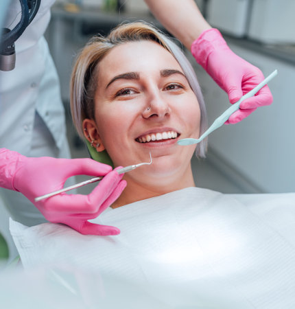 Portrait of young smiling woman sitting in stomatology clinic chair and doctor's hands with dental Mouth mirror and Dental explorer tools preparing to examine teeth condition. Health care concept.の写真素材