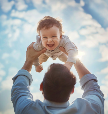 Beautiful little son cheerfully laughing at camera while dad playing rising overhead and lovely smiling to him on blue sky with white clouds background. Family values, small kids, parenthood concept.の素材