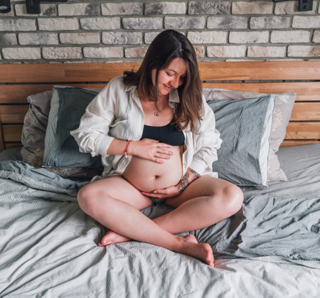 Young pregnant woman sitting with cross-legged tender touching belly on the bed in bedroom in the early morning time. Women's health, happy pregnancy and calm mental mood concept image.の写真素材