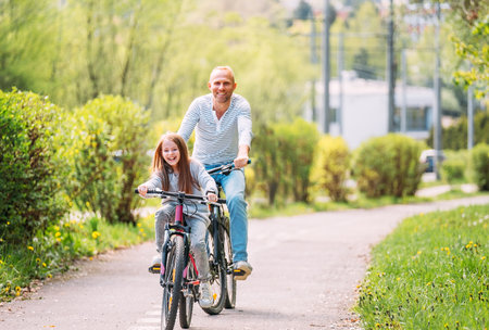 Portraits Smiling father with daughter during summer outdoor bicycle riding. They enjoy togetherness in the summer city park. Happy parenthood and childhood or active sport life concept image.の写真素材