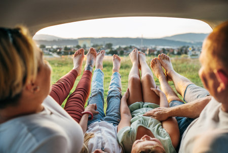 Four-person family lying in the car trunk and fooling around raising feet up. The young caucasian couple has an auto trip break. Family values, traveling, and friendship concept.の写真素材