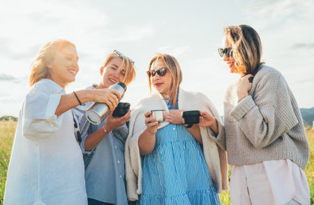 Portrait of four cheerful smiling women drinking tea from thermo flask while they walking by green grass meadow. They looking at the camera. Woman's friendship, relations, and happiness concept imageの写真素材