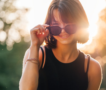 Beautiful young teenage girl in fancy sunglasses dressed in black dress challenging smiling at the camera with a warm sunset backlight. Beautiful people and a fashion concept image.の写真素材
