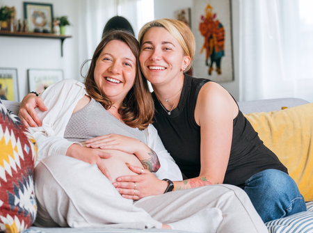 Happy smiling women couple waiting for baby. Same-sex pregnant marriage couple on home living room sofa. Woman's health, happy pregnancy doula supporting and calm mental mood concept imageの写真素材