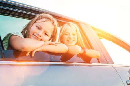 Two happy sisters looking out open car window during auto trip. Cute girls are smiling, laughing during road jorney. Family values, traveling concept side car view photo.の写真素材