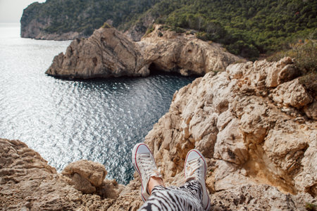 POV shot of female feet in fancy sneakers while Woman sitting on cliff enjoying Mediterranean Sea blue bay waves in morning hours. Mental health, people relaxing, traveling concept, Mallorca, Spain.の写真素材