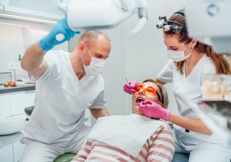 Dental clinic patient appointment. Dentist woman in magnifying glasses doing teeth prevention using medical tools. Young man assistant pointing light. Health care and medicare industry concept image.の写真素材