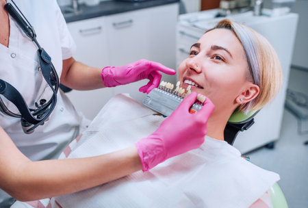 Young female sitting in stomatology clinic chair and smiling showing her teeth. Dentist doctor choosing tooth colors with shade tabs. Healthcare, mouth health and medicare industry concept imageの写真素材