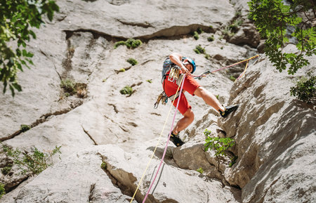 Active climber middle age man in protective helmet while abseiling from cliff rock wall using ropes with a belay device and climbing harness. Active extreme sports time spending conceptの写真素材