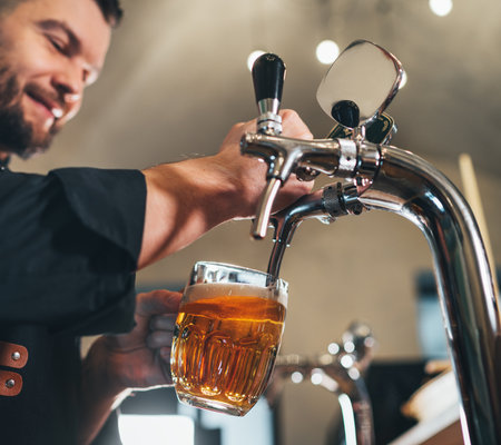 Smiling stylish bearded barman dressed black uniform with an apron tapping fresh lager beer into glass mug at bar counter. Successful people, beer consumption, beverages industry concept imageの写真素材