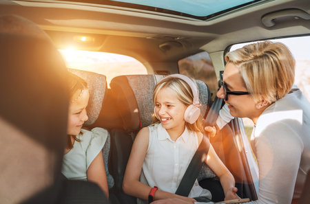 Inside the car photo of a mother fastening with safety auto belt her little daughter girl sitting in child seat. Girl listening music using headphones. Family values, traveling and technology conceptの写真素材