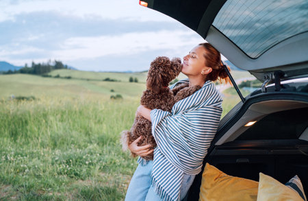 Woman cuddles her brown Maltipoo next to an open car trunk, wrapped in a striped blanket, amidst a picturesque landscape. The scene captures warm, affectionate moment during serene countryside outingの写真素材