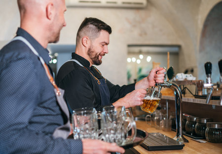 Two caucasian men friends barman beer tapping and waiter with tray dressed black uniform at bar counter. People at work, team work, men friendship and good mood concept image.の写真素材
