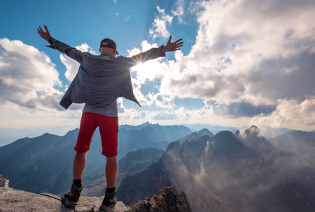 Trekker with raised arms standing on Rysy peak 2499m enjoying High Tatras mountain range covered misty clouds early morning in with incredible light and shadows play. Nature, trekking, climbing concepの写真素材