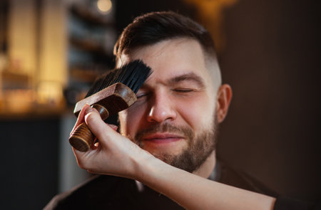 Bearded man portrait in Hair Salon Chair enjoying hairstyle treatment while gently female barber arm cleaning face using neck Brush. Modern low light black styled barber shop interior.Haircare serviceの写真素材