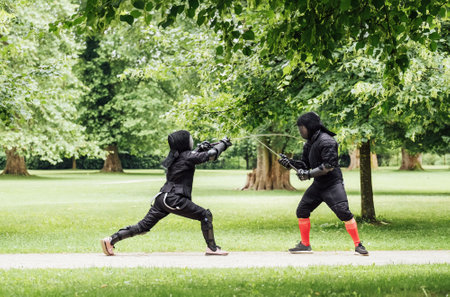 Two fencers dressed in black protective uniforms, helmets with face masks are fencing in city park using long medieval metal historical swords, practicing before competition s. Active people concept.の写真素材