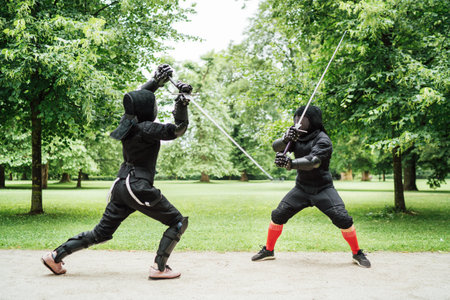 Two fencers dressed in black protective uniforms, helmets with face masks are fencing in city park using long medieval metal historical swords, practicing before competition s. Active people concept.の写真素材