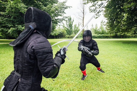 Two fencers dressed in black protective uniforms, helmets with face masks are fencing in city park using long medieval metal historical swords, practicing before competition s. Active people concept.の写真素材