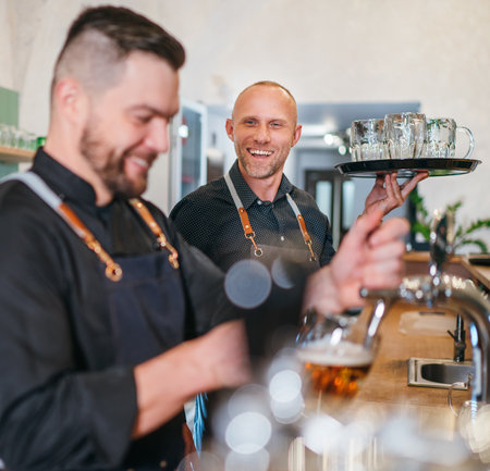 Two men friends barman and waiter with tray dressed black uniform at bar counter chatting smiling to...