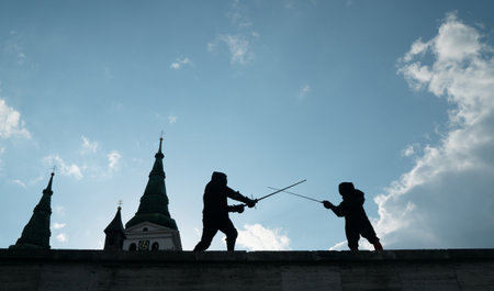 Two fencers dressed in black protective uniforms, helmets with face masks are fencing on castle wall using long medieval metal historical swords practicing before competition s. Active people conceptの写真素材