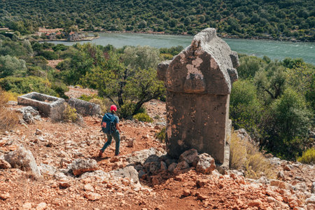Lonely hiker woman walking net to Stone sarcophagus at ancient Simena KalekÃ¶y city in Turkey. Mediterranean sea surrounded by rugged hills listed morning light. Archaeological treasure of Lycia.の写真素材