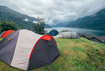 Tents on green hillside overlooking serene fjord with misty mountains in background. Scenic camping...