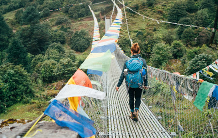 Young female trekker crossing river over Suspension Bridge decorated with multicolored Tibetan Prayer flags hinged over gorge near Lukla. Everest Base Camp route, Sagarmatha National Park, Nepalの写真素材