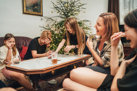 Cheerfully warm home togetherness cozy scene of family playing cards together in home livingroom. Girls smiling and laughing with moms next Christmas tree. Holidays time spending relations conceptの写真素材