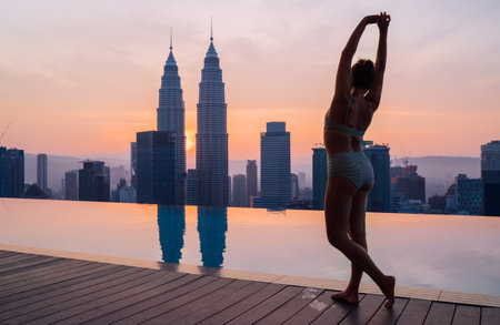 Woman in swimsuit posing for photo next to infinity pool enjoying two skyscrapers tower twins view during evening sunset time. Traveling in Malaysia capital luxury hotel concept imageの写真素材