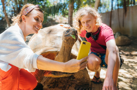 Smiling tourists boy with mom making selfie using cell phone with Aldabra giant tortoise endemic species - one of the largest tortoises in the world in the zoo nature park on Mauritius island.の写真素材