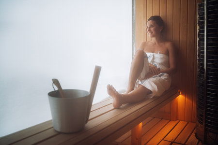 Relaxing Woman wrapped white towel lying on the wooden bench in Hot Finnish sauna with a wide window near hot stones and enjoying pleasant body care temperature treatment. Healthy lifestyle conceptの写真素材