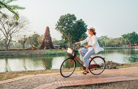 Female traveler in light summer clothes and hat enjoying early morning bicycle ride through Ayutthaya historical park, Thailand. Ancient ruins and serene atmosphere create culture and tranquility vibeの写真素材