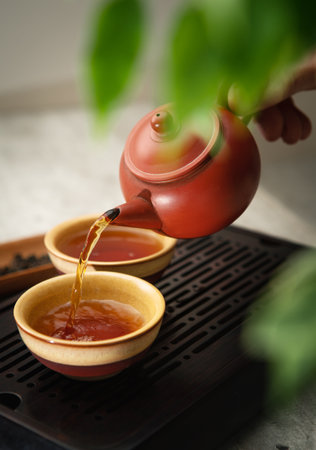 Female hands pouring tea from yixing clay teapot into ceramic tea cups, creating serene tea ritual. Soft light, wooden tea tray and greenery evoke calming mindful ceremony atmosphere.の写真素材
