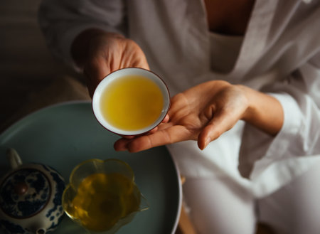 Hands holding porcelain teacup filled with golden tea, exuding warmth and tranquility. Elegant setup with teapot and glass pitcher enhances an intimate, soothing tea ceremony experience.の写真素材