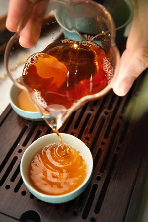Female hands pouring tea from clear glass pitcher teaware into tea cups, creating serene tea ritual. Soft light, wooden tea tray evoke calming mindful ceremony atmosphere.の写真素材