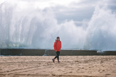 Lonely backpacker woman dressed red softshell jacket enjoying winter storm Atlantic waves crashes on...