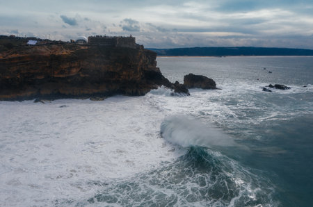 Atlantic ocean waves crashing over rocky coast during international surfers challenge at Farol da NazarÃ© in Nazare, Portugal on Moody cloudy February day.の写真素材