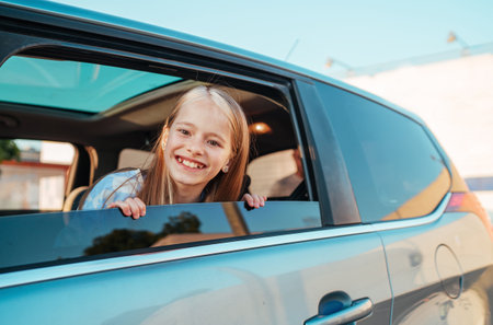 Portrait of smiling little girl looking out from an open car window while she sitting in back seat during family car journey. Traveling by car, traffic safety and modern family concept.の写真素材