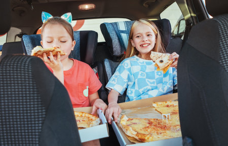 Portrait of two positive smiling sisters eating italian pizza sitting in child car seats on car back seat.の写真素材