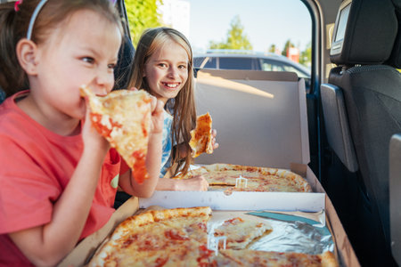 Portrait of two positive smiling sisters eating italian pizza sitting in child car seats on car back seat. Happy childhood, eating or auto journey lunch break concept image.の写真素材