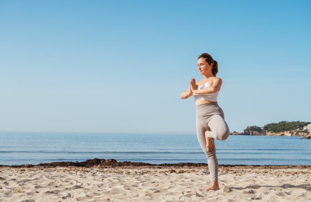 Calm woman standing on one leg doing deep breathing and stretching exercises and meditating in early morning hours on sandy beach with calm sea waves. Mental health, people relaxing, traveling conceptの写真素材
