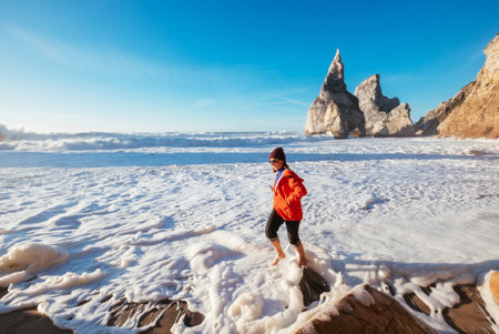 Lonely woman dressed red jacket walking by Beautiful abandoned Atlantic ocean Ursa beach and enjoying coastal surf with foamy waves crashing on rocky cliffs on Winter February coast in Portugal.の写真素材