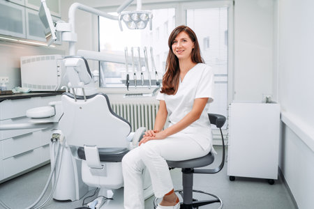 Portrait of sincerely smiling young dentist woman dressed white medical scrubs uniform sitting in modern dental clinic next to stomatology chair. Health care and medicare industry concept image.の写真素材