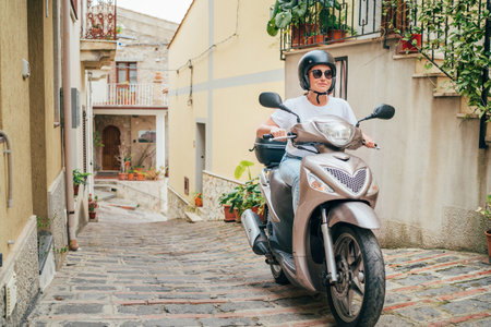 Cheerfully smiling woman in helmet and sunglasses fast riding the moto scooter on the narrow Sicilian old town streets. Happy Italian vacation and transportation concept.の写真素材
