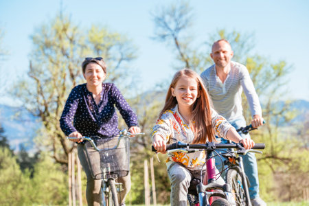 Smiling father and mother with daughter during summer outdoor bicycle riding. They enjoy togetherness on green high grass meadow. Happy parenthood and childhood or active sport life concept imageの写真素材
