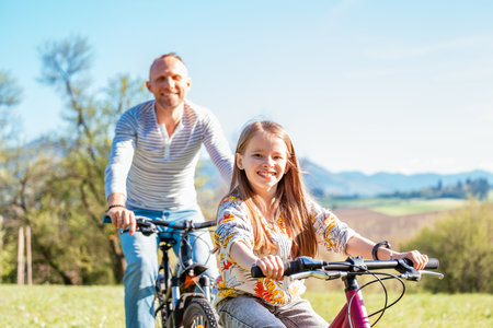Portrait of a smiling father with his daughter during a summer bicycle ride outdoors. They enjoy quality time together in a sunny city park. Happy parenthood, childhood, and active lifestyle concept iの写真素材