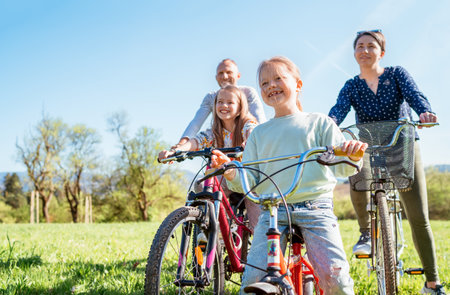 Smiling father and mother with their two daughters during summer bicycle ride outdoors. They enjoy quality time together in a sunny city park. Happy parenthood, childhood, and active lifestyle conceptの写真素材