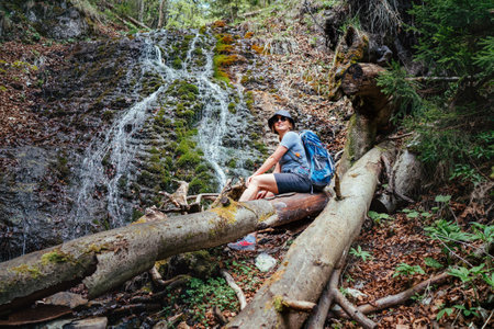 Smiling woman with backpack in hat dressed in active trekking clothes and boots sitting near mountain river waterfall and enjoying splashing Nature power. Traveling, trekking, nature concept imageの写真素材